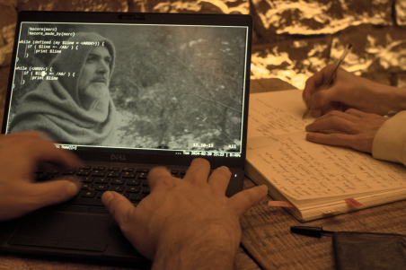 Un ordinateur portable posé sur une table avec deux mains tapant au clavier. Sur l’écran, un éditeur de texte avec une image du film « Le Nom de la Rose ». À côté, deux mains écrivant sur un vieux livre.